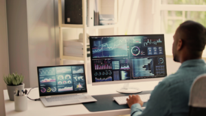 A person sits at a desk with a laptop and large monitor displaying various charts, graphs, and data visualizations crucial for their research proposal. A potted plant adds a touch of green, while shelves bask in the sunlight in the background.